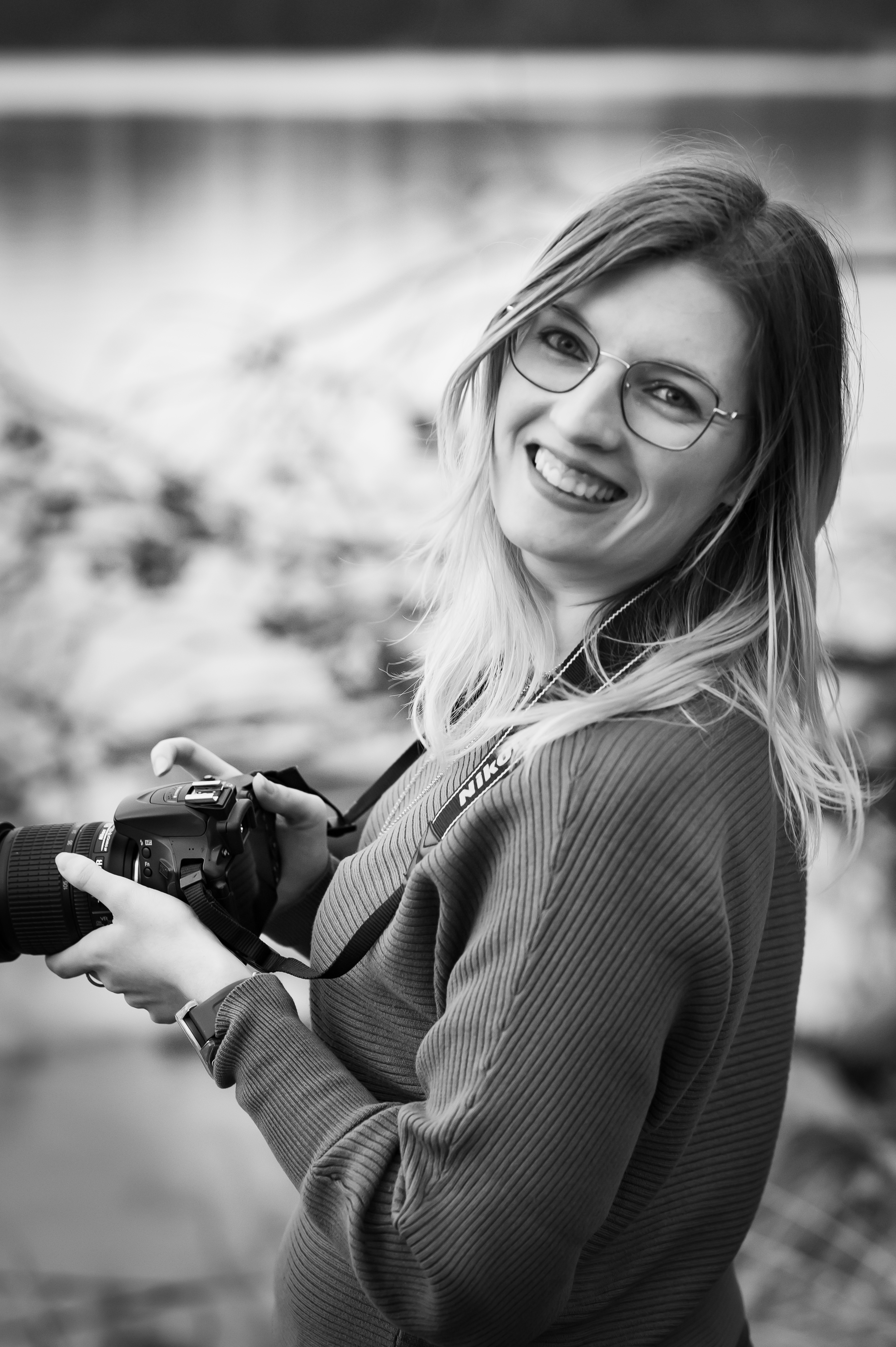 Fotografin Caroline Nagl (Carografie) in schwarz weiß mit Kamera und Lächeln bei der Arbeit in Linz am Weikerlsee